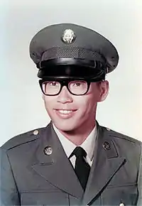 Head and shoulders of a young man wearing a peaked cap, black thick-rimmed glasses, and a military jacket with a round pin on each lapel over a shirt and tie