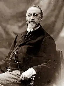 seated middle-aged white man, with medium-length moustache and neat beard, looking benignly towards the camera