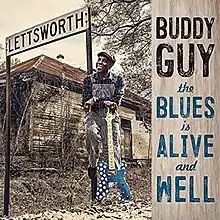 Buddy Guy, wearing overalls and holding an electric guitar, stands near a sign saying "Lettsworth"