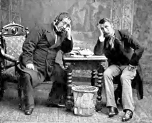 Two youngish men in 19th century morning dress, sitting on either side of a small table, looking baffled