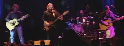 Four band members are performing. Koppes is facing forward and strums his guitar. Kilbey is playing a bass guitar and singing into a microphone. Powles is set back, obscured by his drum kit. Willson-Piper is partly turned to his left and is strumming a guitar.