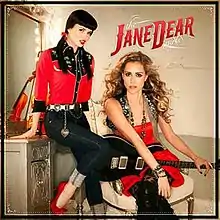 An image of two women looking toward the camera; one is wearing a red dress, while the other is wearing a red shirt and jeans. The album's title is superimposed over the image.