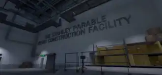 A storage facility with grey floors and off-white walls. Catwalks are suspended above the player, and there are some boxes stacked over to the right. On the wall in block letters is the phrase "The Stanley Parable Demo Construction Facility".
