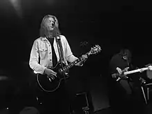 Long haired male singer with Les Paul guitar standing on stage at the microphone in a black and white shot taken from a lower position in the audience