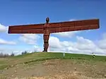 A large rust-coloured statue of a figure stretching wing-like arms out on top of a hill.