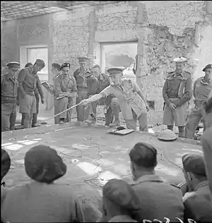 Brigadier A. C. Willison uses a pointer to show the men around him something on a sand table