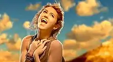 A brunette teen tilts her head upward and looks to the sky while clasping her hands at her chest and singing: She sits at the top of a red mountain cliff during sunset.