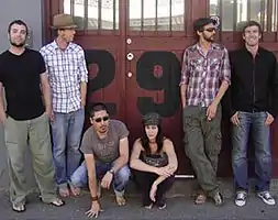 Six people are shown in front of a large building's closed doors. Standing at left is a standing man with dark hair and beard. He wears a black t-shirt and grey pants with his hands tucked in the pockets. He stares forwards. The second standing man has his head turned to his left. His hair is dark and he has a sparse beard. He wears a brown hat, a white shirt with blue squares and jeans with his left hand tucked in. The third man is squatting with his right hand on the surface in front and his left arm resting on his thigh. He has short, dark hair and a trimmed dark beard. He wears a grey-brown t-shirt, sunglasses and jeans. The fourth person sits on the stoop with her elbows at her knees, her right arm holds her left lower arm, which dangles down. She has a grey-black cap over her very long, black hair which is draped over the front of her shoulders. Her top is grey and pants are black. The fourth man is standing and also has his head turned to his left. His right leg is crossed at the ankles. He wears a dark beret, a red, white and black checked shirt over dark, grey pants. He has dark hair and beard. His right arm is tucked in, while his left holds a cigarette. The last man is clean shaven, with dark hair, and he smiles broadly. He wears a black zipped up jacket over his jeans with his thumbs tucked into the pockets. The brown doors include barred windows and have black numbers printed below the windows: only "2" and "9" are visible.