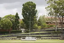 A grassy playing field with a war memorial at the end. Above the memorial are terraced steps leading up to trees and a chapel.