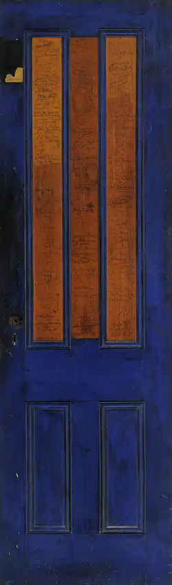Image of a thin door painted blue except for two rectangles in the middle. There are ink signatures covering it.