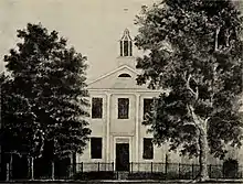 Drawing of a two-story white building with a cupola on the roof upon which is a cross. It is behind a fence and surrounded by two large trees.