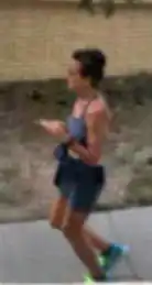 A woman with short brown hair in running motion, wearing a gray tank top and darker gray tight-fitting shorts and neon blue and green running shoes. She is on a sidewalk with a cream-colored wooden fence behind her
