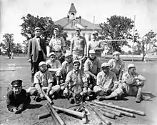 Group photograph of baseball players wearing 1910s-era uniforms, standing, kneeling, or laying on the ground behind a collection of baseball bats