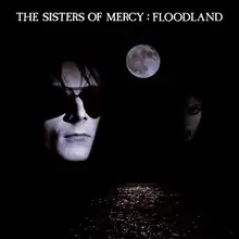 A flooded street at nighttime, with the Moon visible in the sky and two faces peering out of shadows in the background. In white text, "The Sisters of Mercy: Floodland" is written in all capitals at the top of the picture.