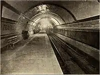 A circular tunnel with platform on the left and tracks on the right. The platform wall is tiled in bands with the words TRAFALGAR SQUARE picked out in coloured tiles