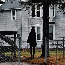 A photograph of a woman dressed in black standing with her back to the camera, facing a run-down house. No words are printed on the cover.