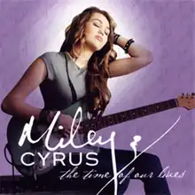 The right profile of a long-haired brunette teenager holding a green and white colored, electric guitar with her face tilting up. She is wearing a gray T-shirt and blue zebra-patterned pants. Beneath her are letter stating in cursive "Miley Cyrus" and "The Time of Our Lives". The background is purple and has cursive writing on it.