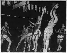 Newspaper photograph of an American Basketball League game between the Scranton Miners and the Elmira Colonels. Leroy Chollet is jumping with one hand raised to contest a jump shot from Danny Finn who is fading away with the ball raised above his head. Three other players position themselves for a rebound near the basket.