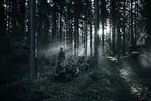 A shot from the film where Finnish soldiers are sitting in a forest whilst the sun shines through between the trees, silhouetting the soldiers.