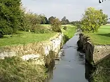 The best preserved of the lock chambers at Alvingham, the retaining walls of brick and stone are intact, but there are no gates. We are looking downstream, away from the weir formed by the upstream sill, which is not visible. On either side of the channel are flat green meadows, and framing those to the edges of the picture are dense woodland.  It is a bright sunny day and the trees and grass are a bright green.  The data for the picture say it was taken in October 2007, but the vivid green of most of the trees, and particularly one just on the right of the lock, looks like spring.