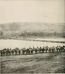 Sepia photograph shows a column of soldiers on horseback with a pontoon bridge in the background.