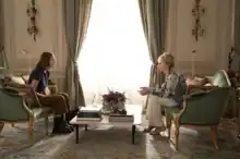 The screenshot is a wide shot of a lavish living room where Ava Daniels and Deborah Vance are seated opposite each other on identical green couches separated by a marble coffee table. The shot shows their profiles.