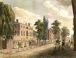 A painting of a sunlit, tree-lined street, with two prominent buildings, each behind a full-height brick wall. Passers-by include women with parasols, a man in a top hat and yellow suit, and a solder in a blue coat with red trim and a white sash.