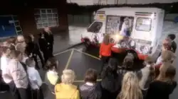 A white ice cream van is parked on a car park in front of a building. A man in a visor and a white jacket is hanging out of a side window of the van, and is handing a glowing object to a young girl in a red jumper and black skirt. Twenty-three similarly aged children stand in a horseshoe shape around the girl.