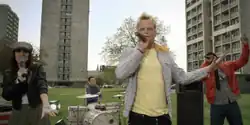 A young woman in a dark leather jacket, a man in a blue jumper, a man with bleached blonde hair in a yellow T-shirt and a man in a red leather jacket are performing on a grassy area outside a tower block estate during daytime. The man in the blue jumper is drumming at a drum kit; the other three are singing into microphones.