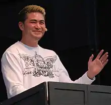 A smiling young man of white, Korean, and Filipino descent, standing at a podium, gesturing with one hand.