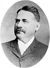 Black and white photo of a middle-aged man with a dark-haired moustache, wearing a suit jacket and white dress shirt