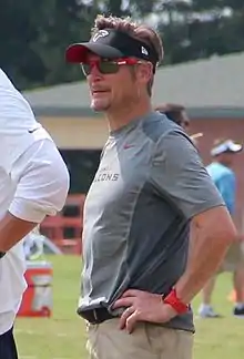 Candid waist-up photograph of Dimitroff wearing a grey t-shirt and black visor both bearing Atlanta Falcons logos and standing on a football practice field with arms akimbo