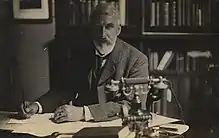 Black and white photo of Barker sitting at a desk with pen in hand and a telephone in the foreground.