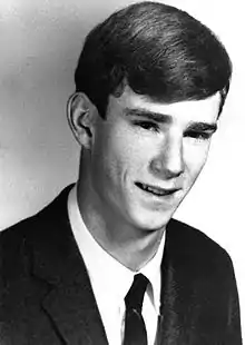 A black and white headshot of a young McMahon wearing a suit and tie. He is turned slightly to the right with his head down and he is smiling.