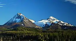 Two volcanoes rise above a landscape of mixed forest and lava, with a glacier spanning the area between the two peaks