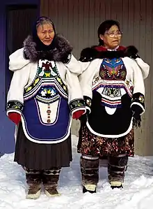 Two Inuit women in clothing covered with detailed beadwork and colorful fabric designs. They have fur ruffs on the hoods of their parkas, cloth skirts beneath their parkas, and fur boots.