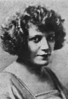 A young white woman with voluminous bobbed curly hair, wearing a dress with a square neckline