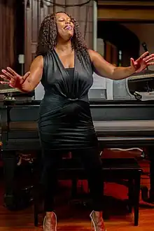A picture of Jackson in a dress, arms stretched out, smiling, at a church, standing behind a piano.