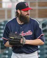 A man in a navy blue baseball jersey, gray pants, and a navy cap with hands held together in his black glove.