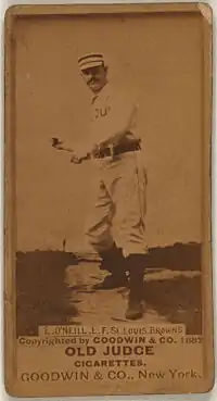 A man in a striped cap, white baseball uniform with the words "ST. LOUIS" obscured on the front and black belt pretends to swing an imaginary bat.