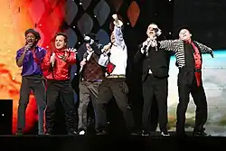 Six men dressed in variously colored clothes performing on a stage.