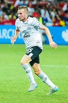 A young man wearing a white top, black shorts, white socks and white boots, standing on a grass field.