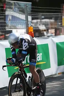 A road racing cyclist in a black and white skinsuit with blue trim and an aerodynamic helmet sits crouched low on his bicycle. There is a guardrail in the background.