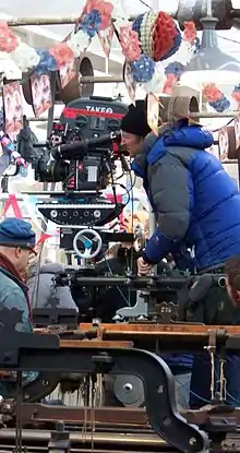  A man wrapped in a blue jacket and black hat looks down the scope of a large film camera. There is red, white, and blue bunting hanging overhead.