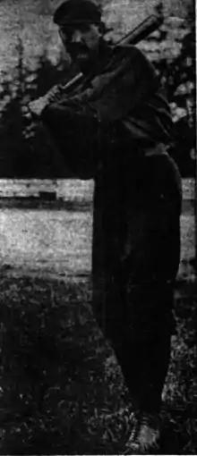 A man with a large mustache wearing a dark baseball uniform and cap stands poised–swing his bat.