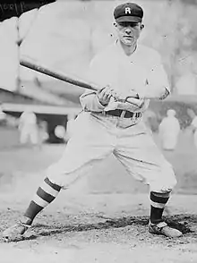 A man in a white baseball uniform with dark pinstripes, high dark socks, and a dark cap with a "R" on the front stands leaning–the right with his bat out–his left side.