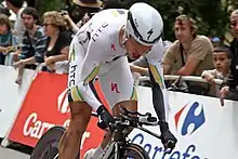 A road racing cyclist, wearing a predominantly white skinsuit with black, green, and gold trim, and an aerodynamic helmet. He is crouched low, but riding out of the saddle. Spectators watch from behind roadside barricades.