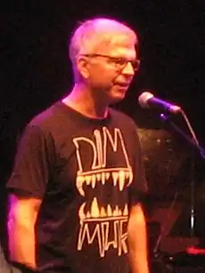 A gray-haired man with glasses and a black shirt standing in front of a microphone