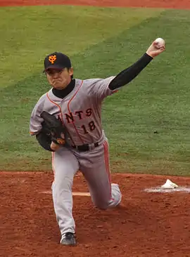 Toshiya Sugiuchi wearing a gray baseball uniform and black baseball cap in the process of pitching a baseball