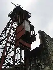 A dark angular structure viewed from its base upwards fills the scene. The sky appears light-grey and cloudy. The structure is made of a dark metal frame surmounted by platform.